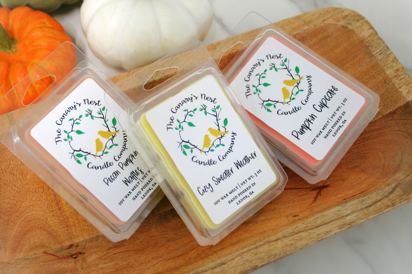 Packaged soy wax melts with fall-themed labels, displayed on a wooden board, with decorative items in the background.